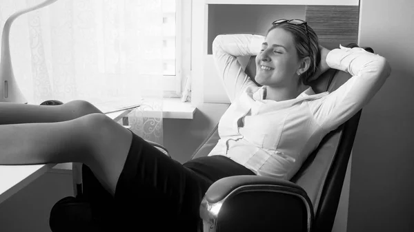 Black and white portrait of smiling young woman sitting in chair and putting feet on office table — Stock Photo, Image