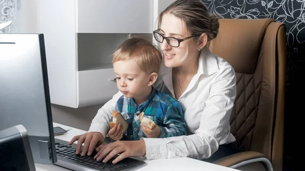 Ładny toddler chłopiec siedzi na kolanach matek w biurze i jedzenie — Zdjęcie stockowe