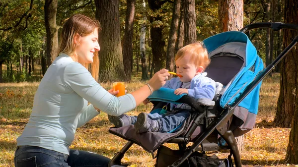 Potret wanita muda tersenyum memberi makan bayinya sambil duduk di kereta bayi dengan bubur — Stok Foto