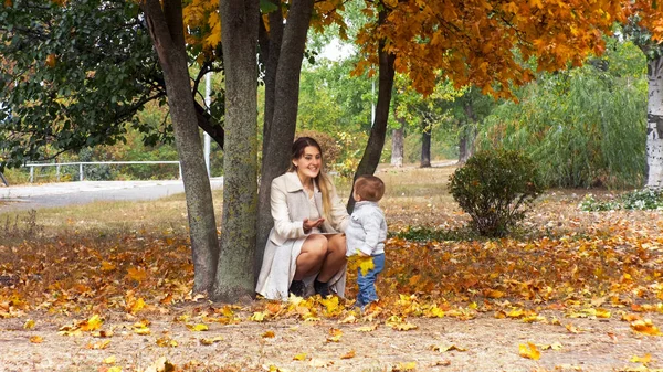 Gülümseyen annesiyle mutlu sonbahar Park ağacın altında oturan yürümeye başlayan çocuk — Stok fotoğraf