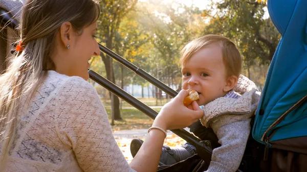 Retrato de jovem mãe dando comida para seu filho bebê faminto sentado no carrinho de bebê no parque — Fotografia de Stock