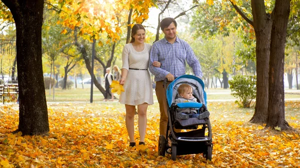 Happy smiling family walking with toddler son sitting in pram at autumn park — Stock Photo, Image