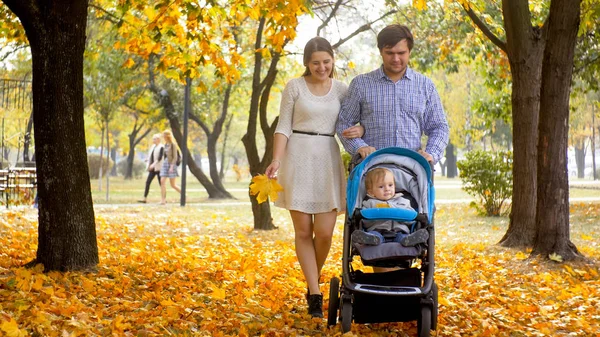 Glada unga föräldrar går med 1-årig baby son sitter i barnvagn på hösten park — Stockfoto