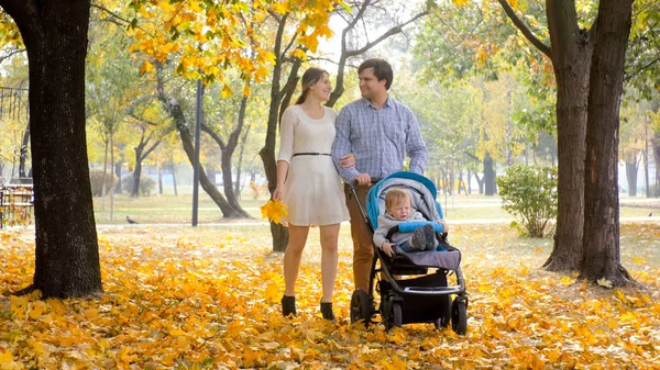 Happy smiling family with baby boy boy in pram walking on yellow leaves at autumn park — Stock Photo, Image