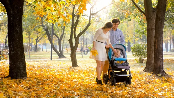 Молодая пара наслаждается прогулкой в осеннем парке со своим годовалым мальчиком, сидящим в коляске — стоковое фото