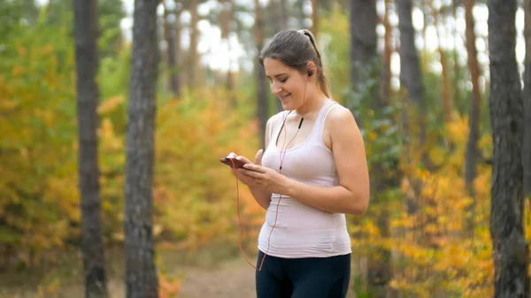Portrait tonique de jeune femme souriante choisissant la musique avant de faire du jogging en forêt — Photo