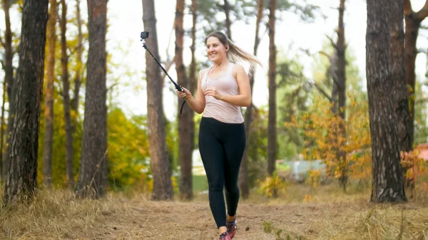 Jovem mulher esportiva correndo com câmera na vara selfie na floresta — Fotografia de Stock