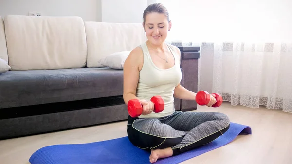 Joven mujer sonriente sentada en una colchoneta de fitness y levantando pesas — Foto de Stock