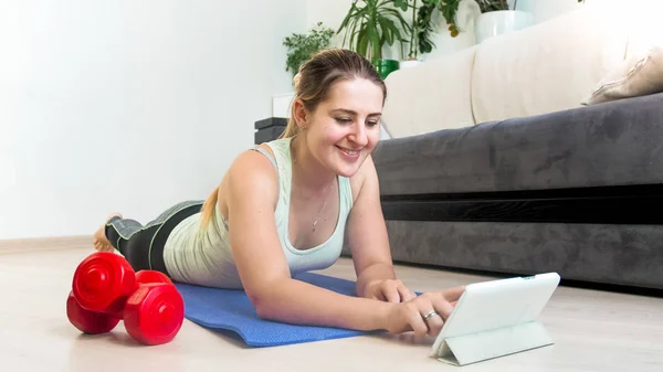 Giovane donna sorridente sdraiata sul pavimento e guardando video trainin su tablet — Foto Stock