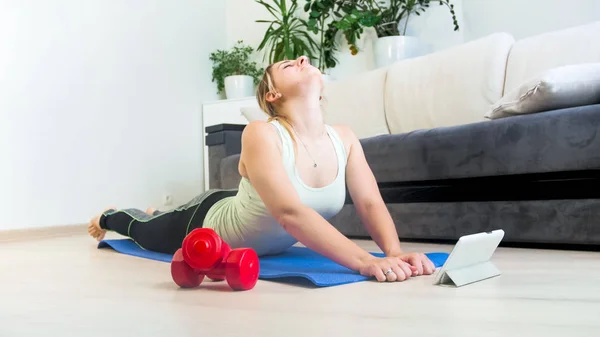 Mujer joven acostada en la esterilla de fitness y escuchando nuevos ejercicios de la tableta digital — Foto de Stock