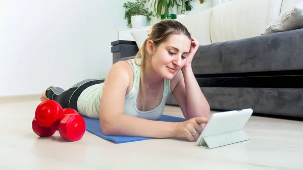 Mujer joven tumbada en el suelo en casa y viendo videos de fitness en la tableta —  Fotos de Stock