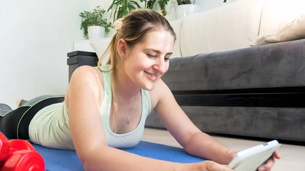 Портрет усміхненої спортивної жінки, що лежить на фітнес-маті і дивиться відео на планшетному ПК — стокове фото