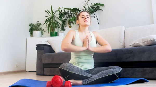 Hermosa joven sentada en una colchoneta de fitness en la sala de estar y practicando yoga — Foto de Stock