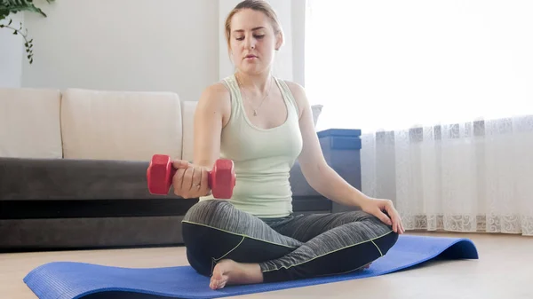 Mujer joven sentada en una colchoneta de fitness en casa y haciendo ejercicio con pesas — Foto de Stock