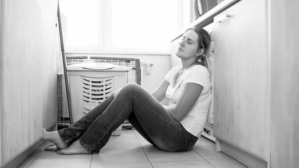 Black and white image of tired woman sitting on floor at laundry room — Stock Photo, Image