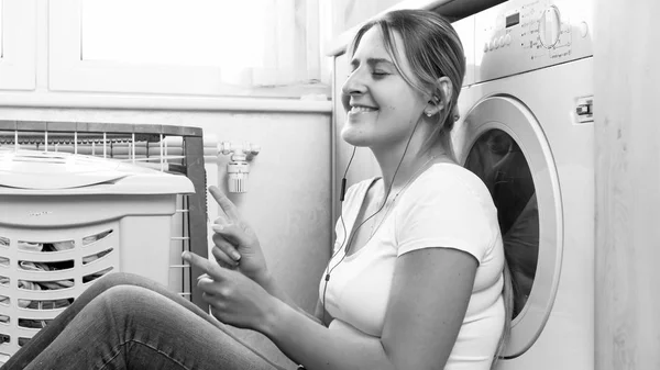 Retrato en blanco y negro de ama de casa sonriente escuchando música con auriculares en la lavandería — Foto de Stock