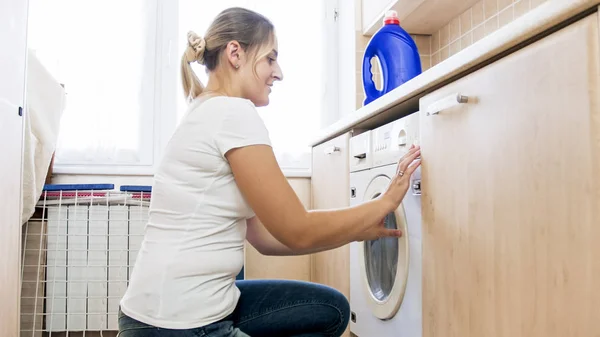 Mulher sorridente bonita fechando porta de vidro na máquina de lavar roupa na lavanderia — Fotografia de Stock