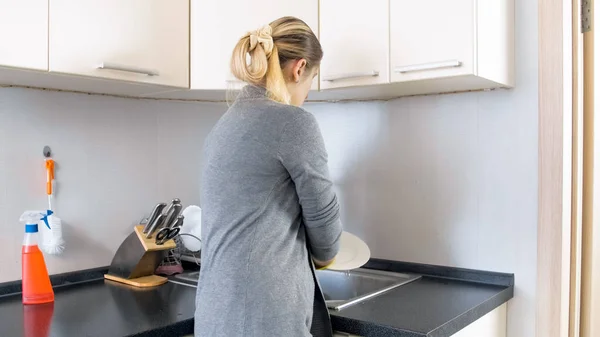 Νεαρή ξανθιά νοικοκυρά πλύσιμο πιάτων στην κουζίνα — Φωτογραφία Αρχείου