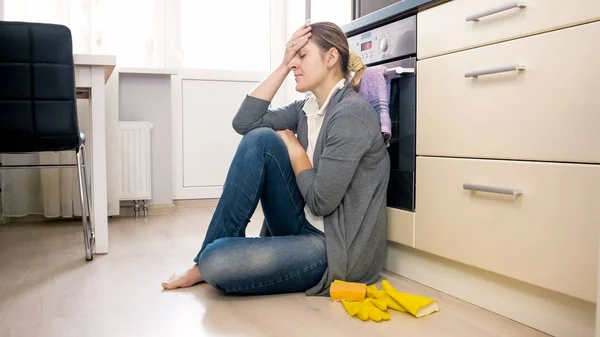 Exhausted woman sitting on floor at kitchen after cleaning house