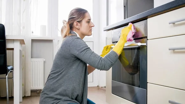 Junge müde Hausfrau in Latex-Handschuhen putzt Ofen in der Küche — Stockfoto