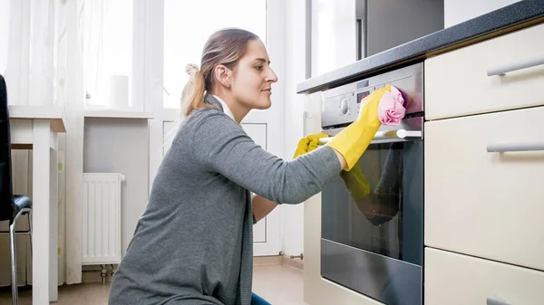 Potret wanita muda tersenyum membersihkan noda dari oven di dapur — Stok Foto