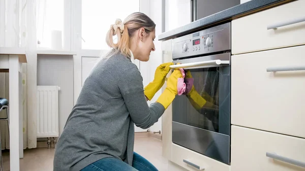 Joven ama de casa en guantes de látex limpiando la puerta de cristal en el horno — Foto de Stock