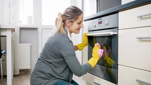 Χαμογελώντας στίλβωσης φούρνο πόρτα γυαλιού νεαρή γυναίκα στην κουζίνα — Φωτογραφία Αρχείου