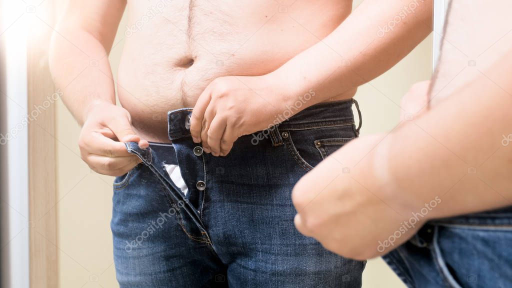 Closeup photo of young fat man buttoning tight jeans at mirror