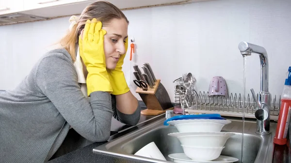 Retrato de ama de casa joven molesta mirando en el fregadero de la cocina con platos sucios — Foto de Stock