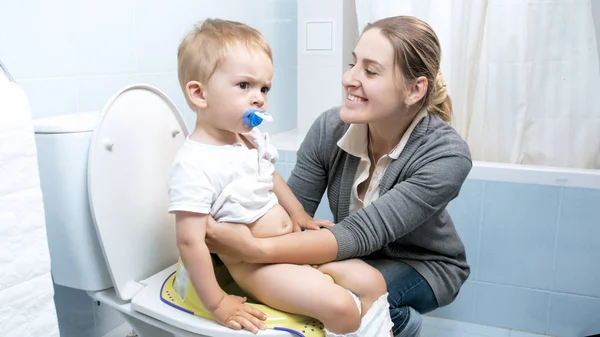 Retrato de la joven madre sonriente sentando a su hijo pequeño en el inodoro — Foto de Stock