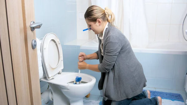 Молодая домохозяйка с прищепкой закрывает нос, чистя туалет дома — стоковое фото