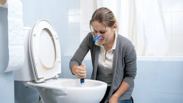 Retrato de ama de casa joven con la nariz cerrada limpieza inodoro sucio en casa —  Fotos de Stock