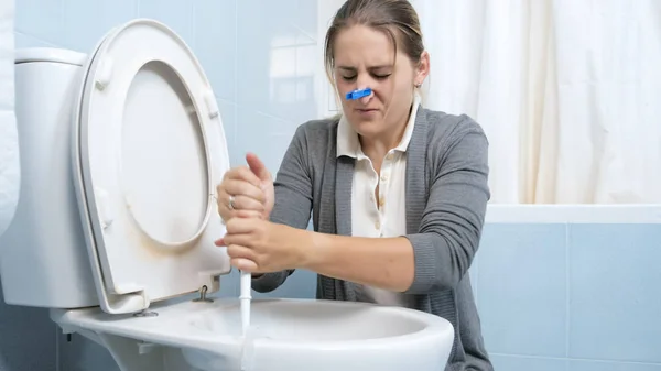 Молодая брезгливая женщина с одеждами на носу, стирающая грязный туалет — стоковое фото