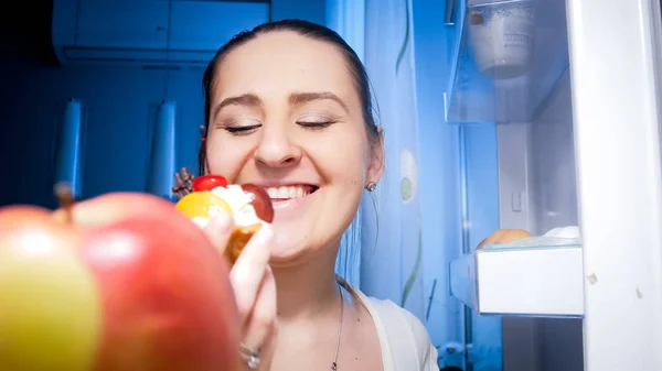 Porträt einer glücklich lächelnden Frau, die nachts süßen Kuchen aus dem Kühlschrank holt — Stockfoto