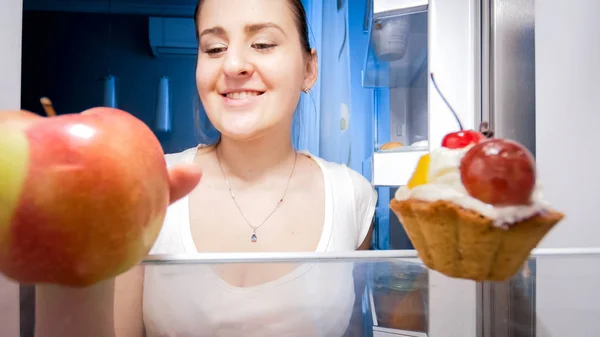 Portrait of young woman taking apple from refrigerator instead of sweet cake — Stock Photo, Image