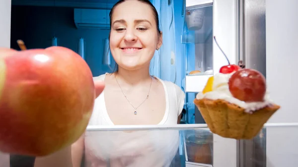 Porträtt av ung leende kvinna tar apple från kylskåp på natten — Stockfoto