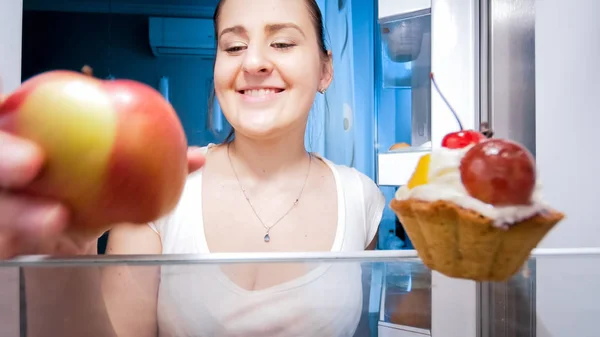 Portret szczęśliwy młoda kobieta biorąc jabłko z lodówki w nocy — Zdjęcie stockowe