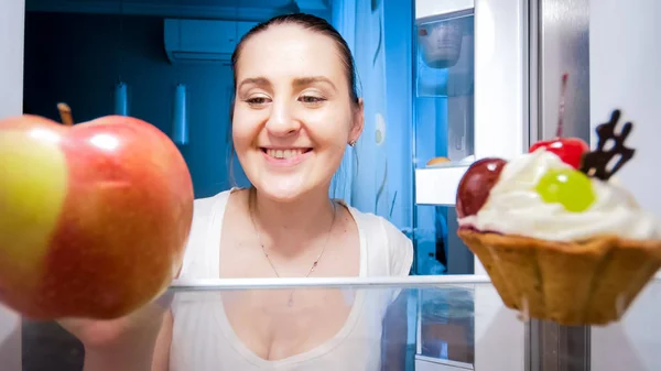 Potret close up dari wanita muda tersenyum meragukan antara makan apel atau kue di malam hari — Stok Foto