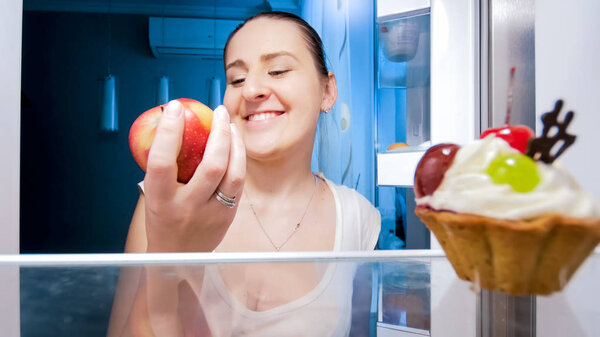Closeup portrait of young smiling woman holding apple on kitchen at night