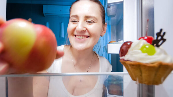 Closeup image of young smiling woman looking inside of refrigerator and taking apple