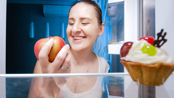 Happy smiling young woman taking red apple from refrigerator at night