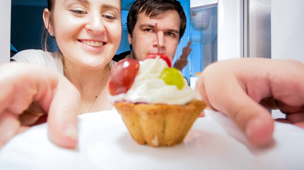 Closeup image of young hungry couple taking sweet cake from refrigerator at night