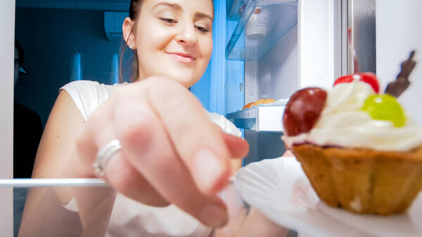Closeup image of young woman taking plate with cake from refrigerator at night
