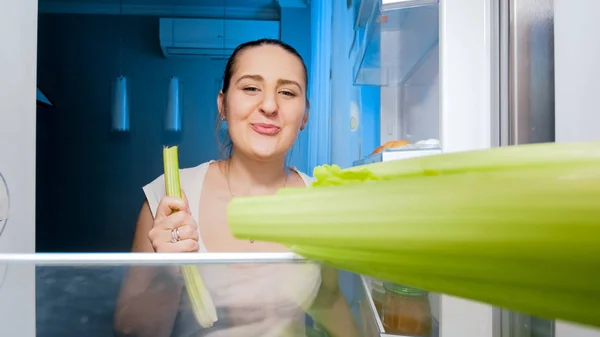 Potret wanita muda tersenyum makan seledri di dapur pada malam hari — Stok Foto