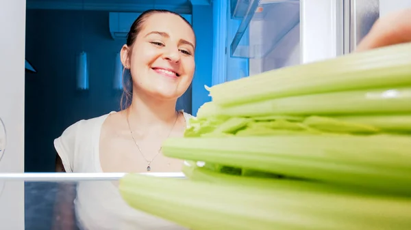 Retrato de una hermosa mujer sonriente sintiéndose hambrienta por la noche tomando apio del refrigerador. Concepto de vegetariano y vegano —  Fotos de Stock