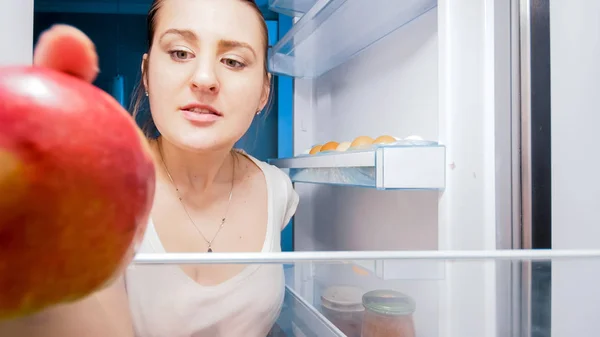 Retrato de cerca de una mujer joven mirando dentro del refrigerador y tomando manzana roja —  Fotos de Stock