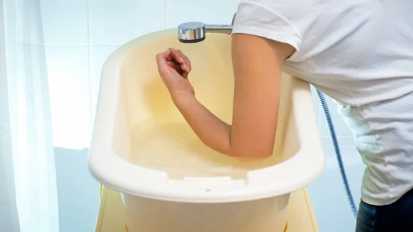 Foto de primer plano de una mujer joven comprobando la temperatura del agua en la bañera del bebé con el codo —  Fotos de Stock