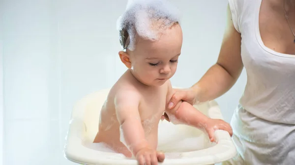 Молодая заботливая мать стирает своего малыша сына в маленькой пластиковой ванне — стоковое фото