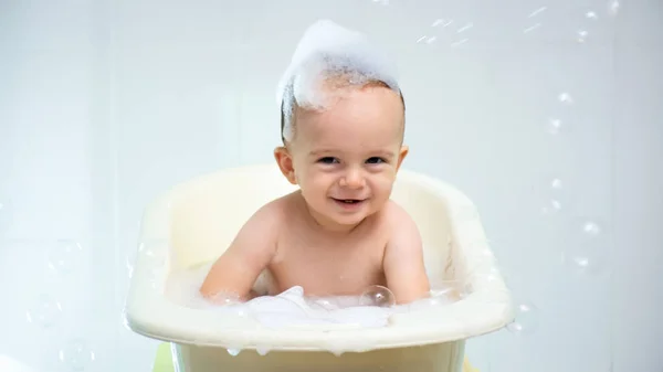 Портрет милого улыбающегося мальчика, сидящего в ванной с кастрюлями на голове и летающими мыльными пузырями — стоковое фото