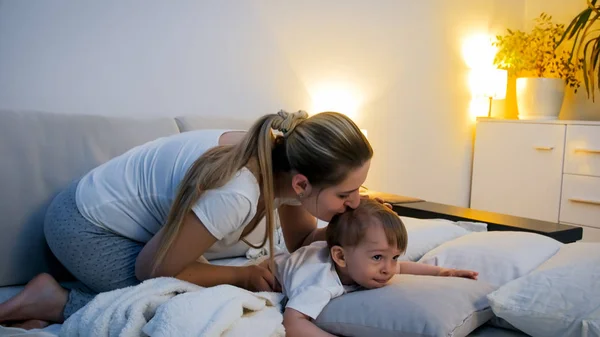 Молодая мать целует своего маленького сына, лежащего ночью в постели — стоковое фото